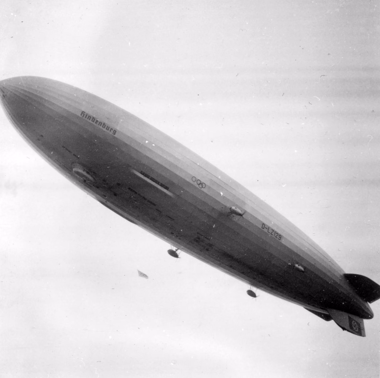 Schwarz-Weiß-Foto der Hindenburg im Flug, schräg von unten gesehen: der lange, silbrige Rumpf mit dem Schriftzug „Hindenburg“, den olympischen Ringen und der Kennung „D-LZ129“, darunter die Motorgondeln, vor hellem, dunstigem Himmel.