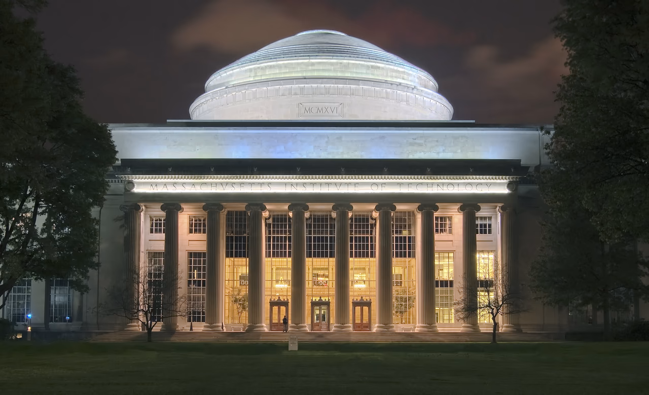 Der Great Dome des MIT bei Nacht: die angestrahlte Kuppel über der Säulenfassade mit erleuchteten Fenstern, davor der dunkle Rasen von Killian Court.