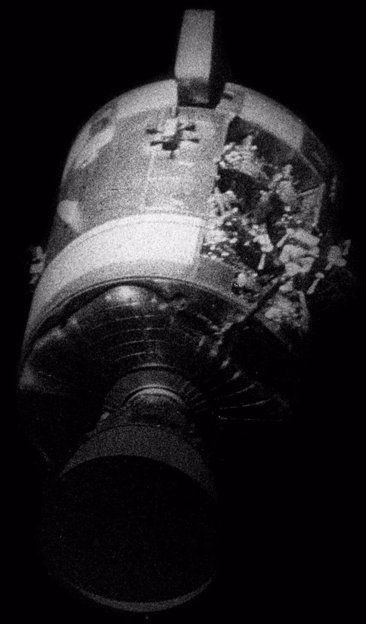 Körnige Schwarz-Weiß-Aufnahme des abgetrennten Servicemoduls von Apollo 13 vor dem schwarzen All: An einer Seite fehlt eine Verkleidungsplatte, darunter liegen zerrissene Leitungen und Geräte frei.