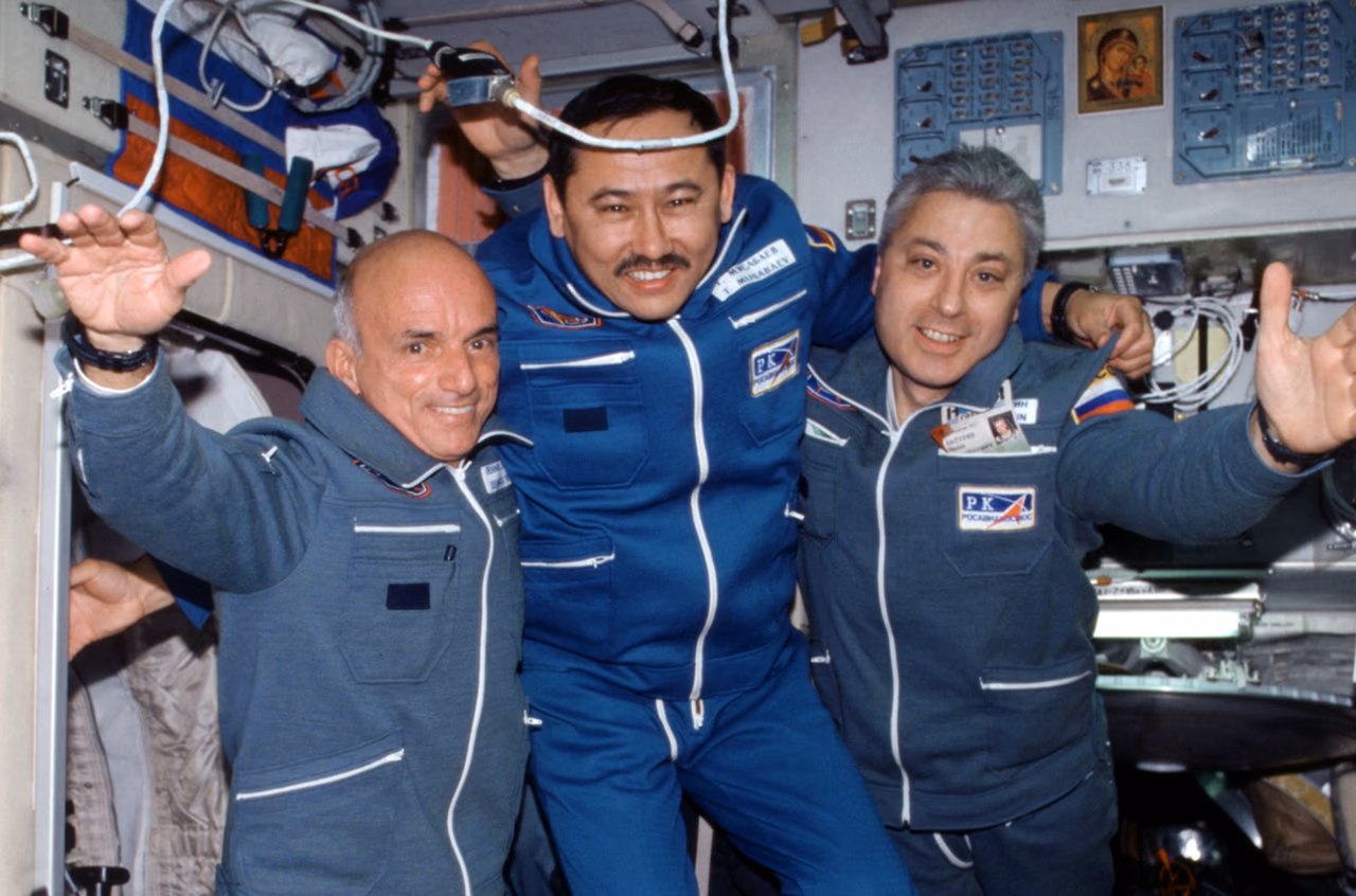 Dennis Tito mit den Kosmonauten Talgat Mussabajew und Juri Baturin in blauen Overalls, schwebend und winkend im Inneren der ISS zwischen Kabeln und Schaltpulten.