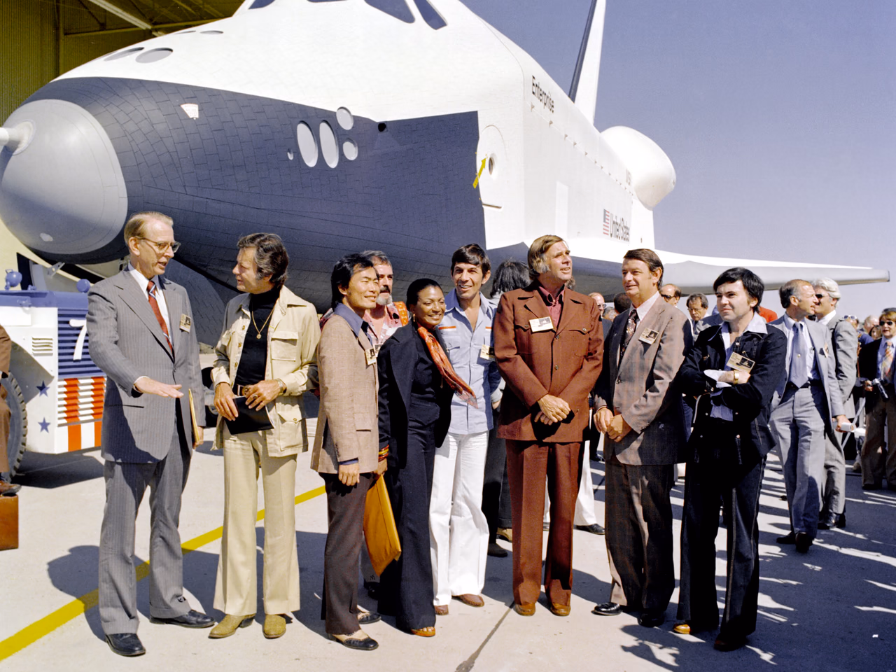 Die Raumfähre Enterprise bei ihrer Vorstellung 1976: Der weiße Orbiter mit dunkler Bugunterseite und dem Schriftzug „Enterprise“ steht vor einer Werkshalle, davor eine Reihe von Gästen in der Kleidung der Siebzigerjahre — unter ihnen mehrere Darsteller aus Star Trek, darunter Leonard Nimoy, Nichelle Nichols und George Takei, sowie der Serienschöpfer Gene Roddenberry.