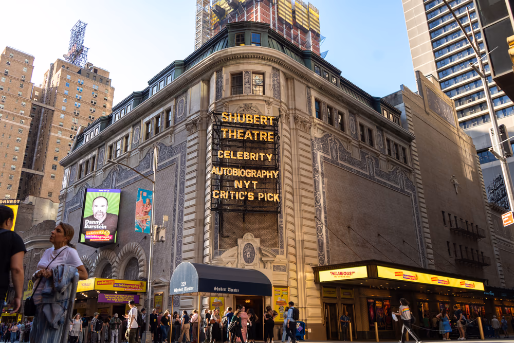 Das Shubert Theatre an einem sonnigen Tag, von der Straßenecke aus gesehen: ein cremefarbener, mehrstöckiger Bau mit abgerundeter Ecke, blaugrauen Ornamentfeldern in der Fassade und einem grünen Kupferdach mit Gauben. An der Ecke hängt eine senkrechte Leuchttafel mit gelben Glühbuchstaben: „SHUBERT THEATRE“, „CELEBRITY AUTOBIOGRAPHY“ und „NYT CRITIC’S PICK“. Darunter führt ein dunkelblaues Vordach mit der Aufschrift „225 Shubert Theatre“ in gebrochener Schrift zum Eingang, davor drängen sich Passanten auf dem Gehweg. Links zeigt ein Werbebildschirm das Porträt eines Mannes mit dem Namen „Danny Burstein“, rechts zieht sich ein gelb leuchtendes Vordach an der Seitenfassade entlang; im Hintergrund ragen Hochhäuser und ein eingerüsteter Neubau in den blauen Himmel.