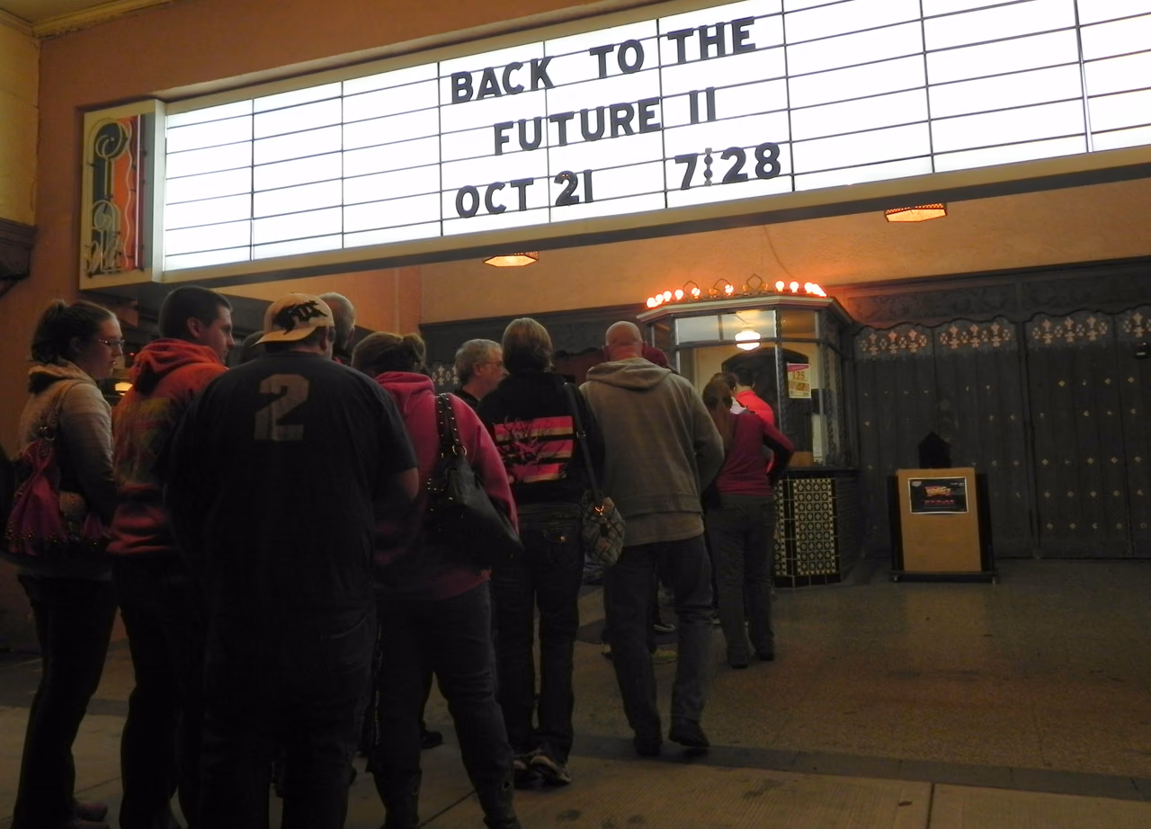 Ein Foto am Abend vor dem Eingang eines alten Kinos: Über einer Menschenschlange leuchtet die weiße Anzeigetafel mit schwarzen Steckbuchstaben „BACK TO THE FUTURE II“, darunter „OCT 21“ und „7:28“. Die Wartenden in Kapuzenpullis und Jacken stehen mit dem Rücken zur Kamera vor einem kleinen Kassenhäuschen, das oben von einer Reihe Glühbirnen gekrönt ist; einer trägt ein Shirt mit einer großen „2“. Links hängt ein buntes Schild im Art-déco-Stil an der Wand. Zu beiden Seiten etwas hinter dem Kassenhäuschen liegen verzierte dunkle Türen, die linken halb verdeckt von den Wartenden, vor den rechten ein hölzerner Aufsteller mit einem kleinen dunklen Aushang, darauf das rot-gelbe englische Logo „BACK TO THE FUTURE II“ und darunter ein unscharfer roter Streifen.